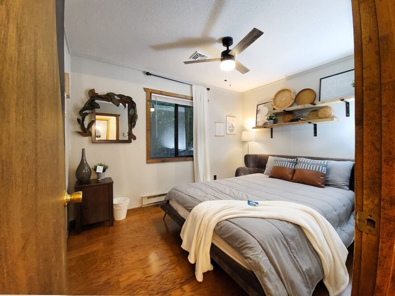 Un dormitorio con cama, ventilador de techo, ventana, mesita de noche y estante con objetos decorativos.