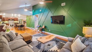 A modern living room with a green accent wall, a flat-screen TV, and a dining area with wooden furniture. - Hot Tub, Game Room, BBQ & Playhouse Oasis – Family Retreat, 5mins to Lake! (Hawley)
