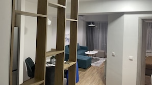 A modern living room with a sofa, a small table, and a chair. There is a wooden partition with shelves, and a ceiling light is visible.