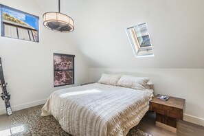 A bedroom with a bed, a small bedside table, a hanging light fixture, a window with a view, and a framed picture on the wall.