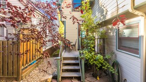 Una casa con terraza de madera, un juego de escalones que lleva a una puerta y un jardín con manguera y planta en maceta.