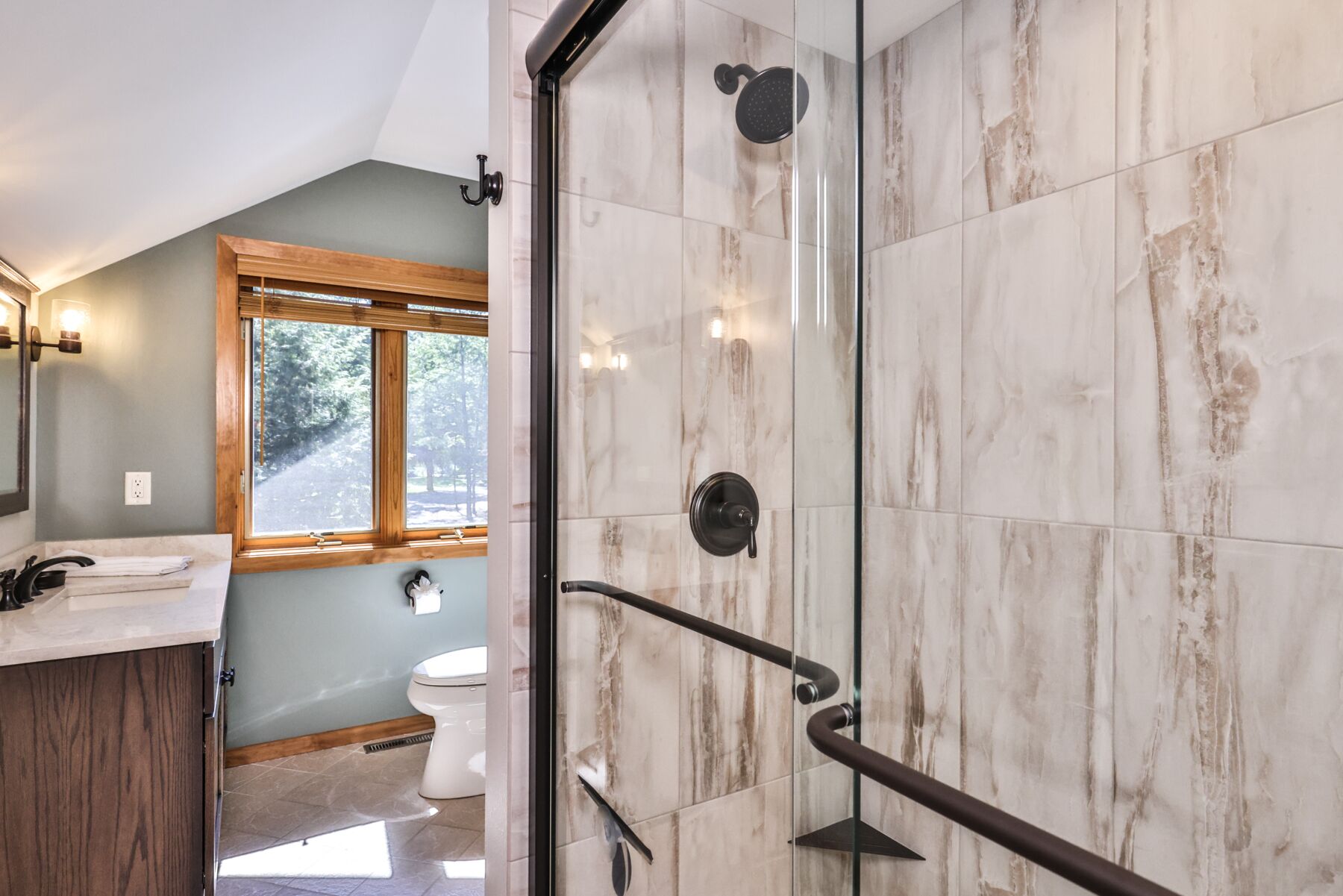 Un baño con cabina de ducha de vidrio, tocador de madera, inodoro y ventana con vistas a árboles.