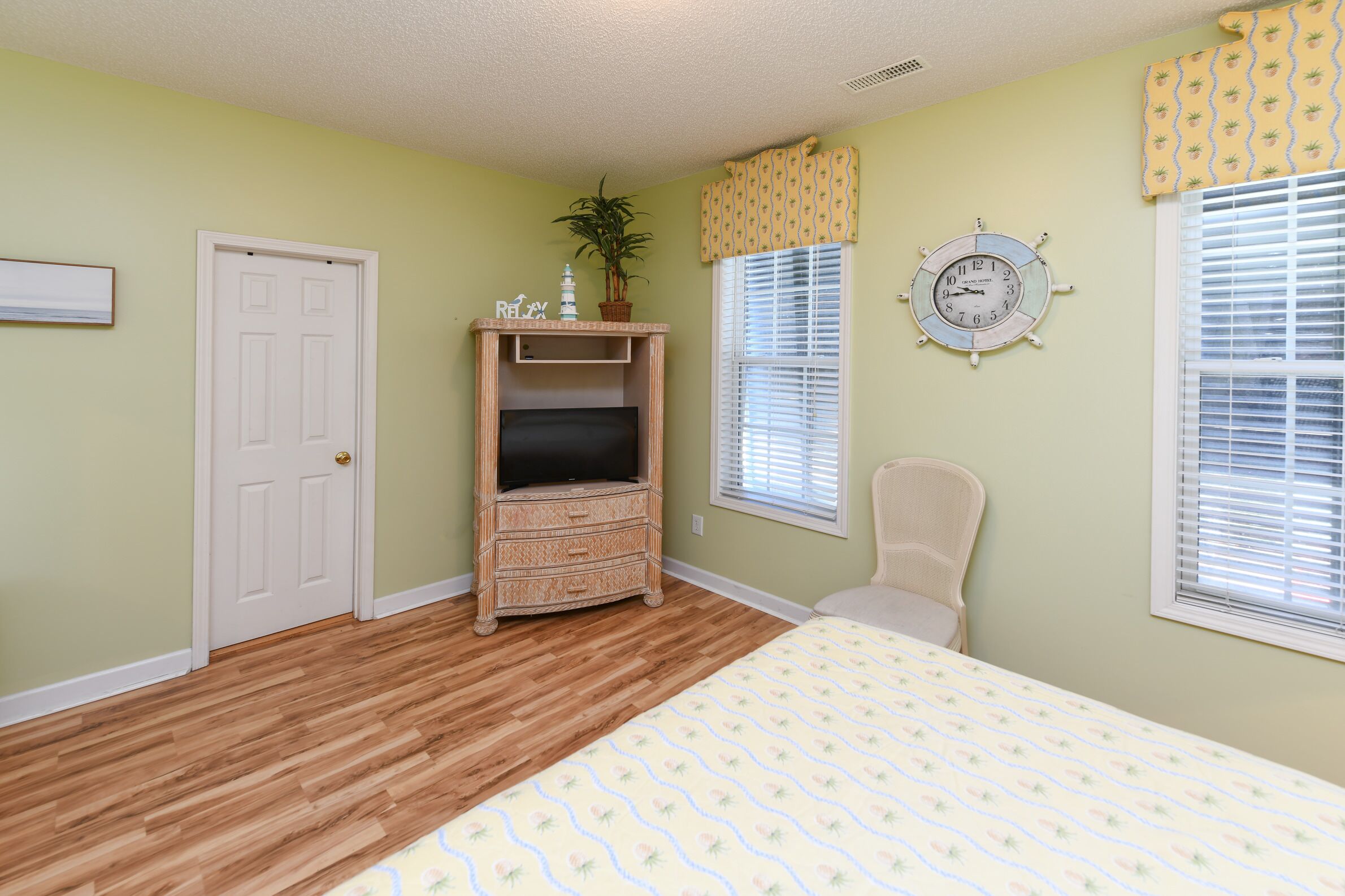 Une chambre avec un plancher en bois, un lit recouvert d’une couvre-lit à motifs floraux, une commode en osier, une chaise, une plante en pot, une horloge murale et une fenêtre avec des stores.