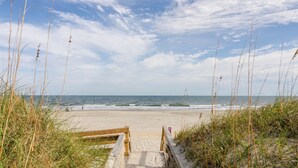 Una spiaggia con un sentiero di legno che conduce alla riva, circondata da erba alta e un cielo sereno.