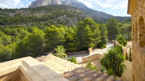 Vista desde un balcón con paisajes montañosos, una cumbre destacada, abundante vegetación y un edificio de piedra con arcos.
