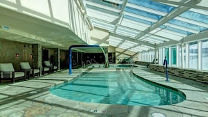 En inomhuspool med ett glastak, stenbelagt golv och solstolar längs väggarna.