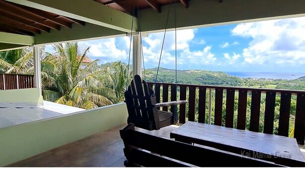 Un balcón con una mesa y sillas de madera, con vistas a un paisaje exuberante con palmeras y vista al océano.
