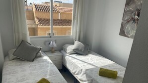 Ein Zimmer mit zwei Betten, jedes mit einem Kopfkissen und einem zusammengerollten Handtuch. Dazwischen steht ein Nachttisch mit einer Lampe.