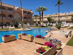 A swimming pool surrounded by flower planters and palm trees, with a building in the background. - Well Equipped 2 Bed Penthouse with Solarium, close to beach and local amenities (Los Alcázares)