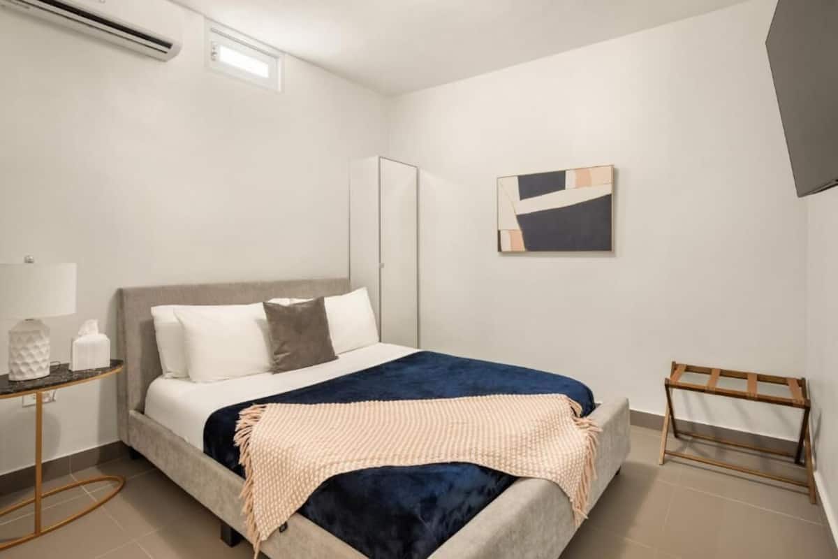 A bedroom with a bed, bedside table, lamp, air conditioning unit, and a wall-mounted artwork.