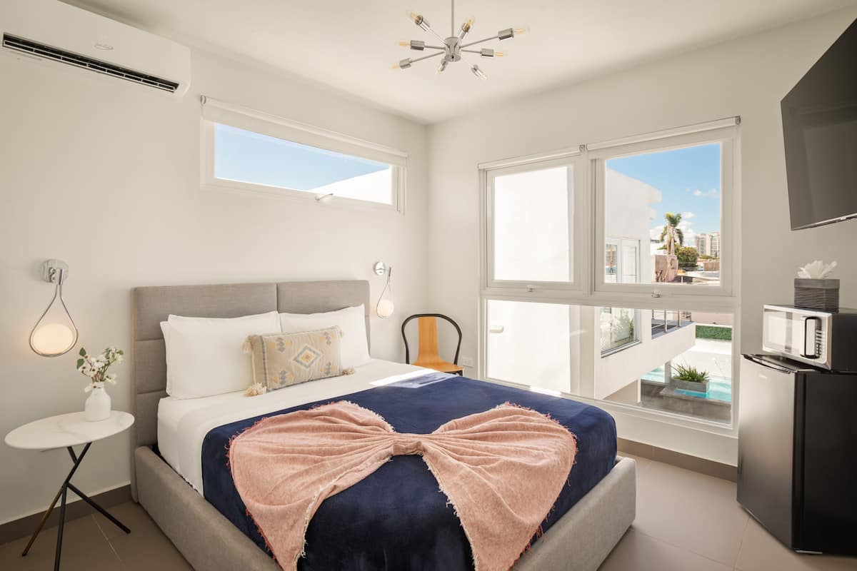 A modern bedroom with a large bed, a small round table, a chair, a TV, and a balcony with a view of buildings.