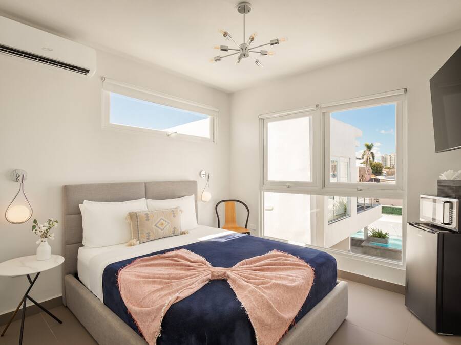 Un dormitorio moderno con una cama grande, una mesita redonda pequeña, una silla, un televisor y un balcón con vistas a edificios.
