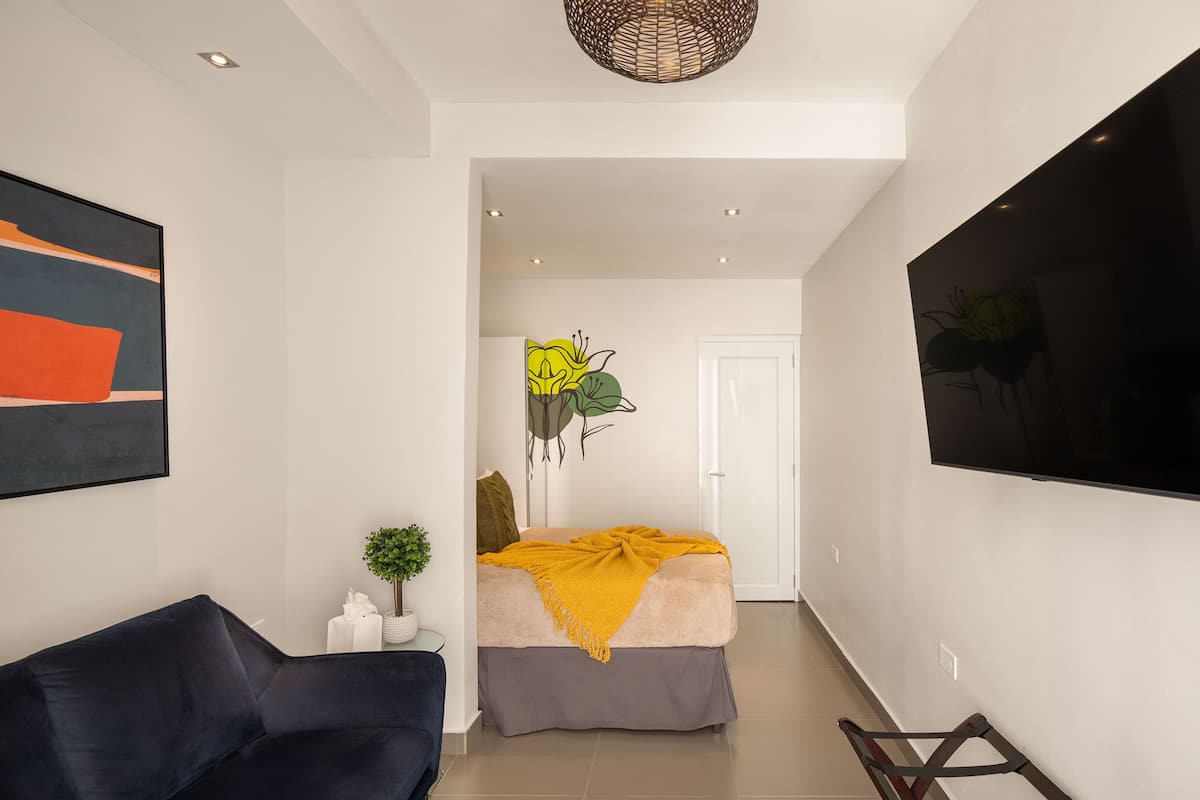 A modern living room with a dark blue sofa, a bed with a yellow blanket, a small plant, and a wall-mounted TV.
