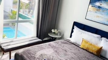 A bedroom with a bed, a bedside table, a window with a view of buildings and a pool, and a painting on the wall.