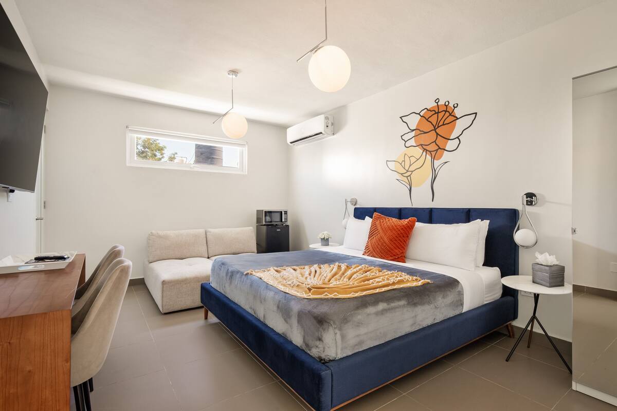 A modern bedroom with a large bed, a desk, and a chair. There is a wall-mounted air conditioner and a small table with a lamp.