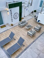 Un área de estar al aire libre moderna con un sofá, sillas y una mesa, rodeada por un edificio blanco con una pared verde y un letrero 'ROSALINA'.