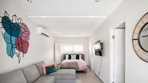 A modern bedroom with a grey sofa, a bed with a grey headboard, a large abstract wall art piece, a round mirror, and a TV on the wall.