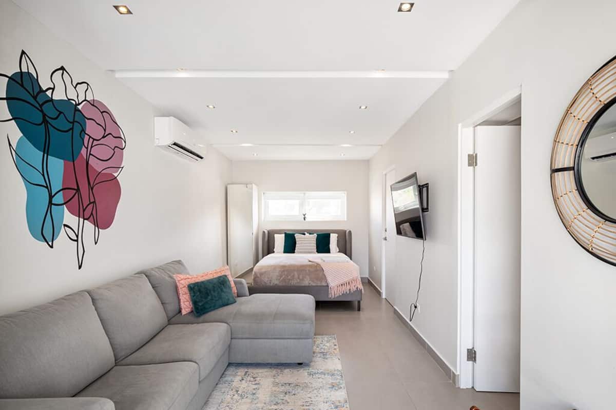 A modern bedroom with a grey sofa, a bed with a grey headboard, a large abstract wall art piece, a round mirror, and a TV on the wall.