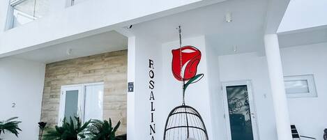 A modern two-story building with a minimalist design, featuring a large flower mural on the facade, a balcony, and a seating area with cushions.