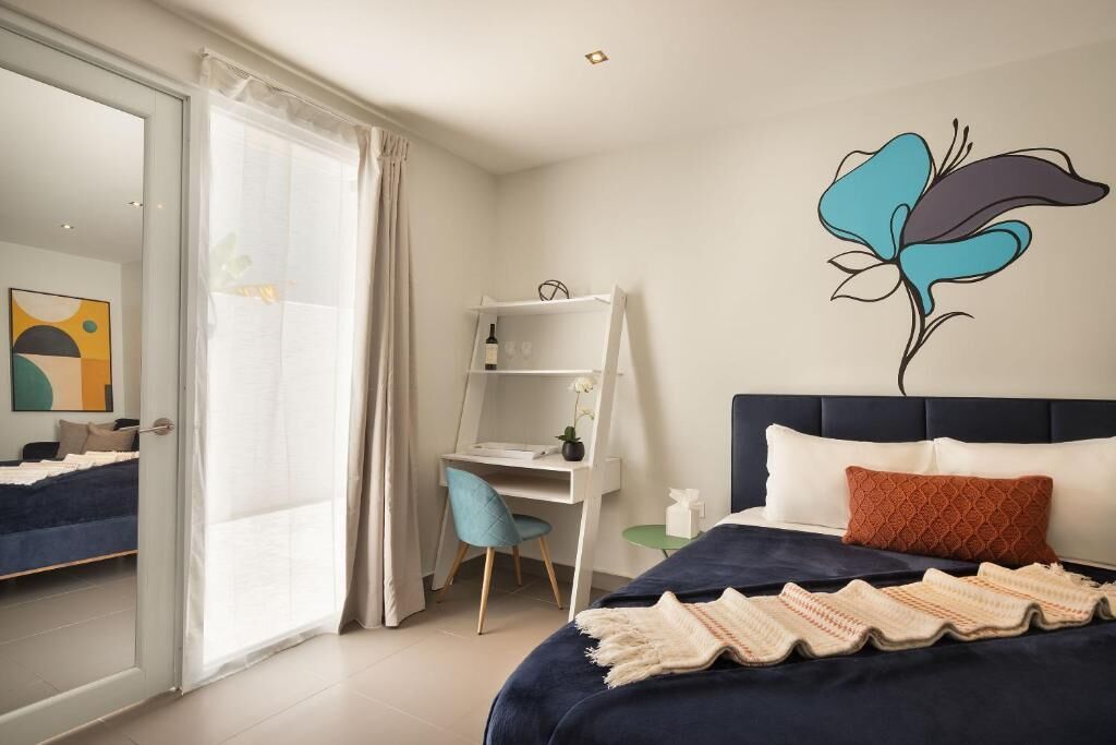 Ein Schlafzimmer mit Bett, Schreibtisch und Stuhl. Es gibt ein Fenster mit Vorhängen, eine Wand mit einer Blumenmotiv-Abbildung und ein Bett mit Decke.