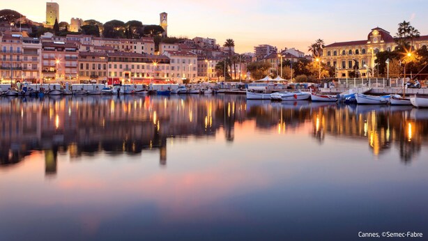 A marina with boats docked, a historic building with a clock tower, and a calm waterfront reflecting the buildings.