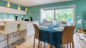 A modern kitchen with white cabinets, a dining area with a blue tablecloth, and a large window offering a view of greenery. - Petite Dépée by Interhome (Grayan et L' Hopital)