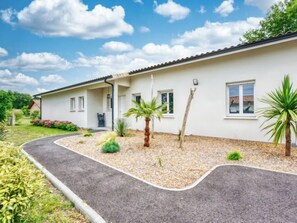 A single-story building with a white exterior and a tiled roof. There are two windows on the side of the building, a door, and a small porch area. The surrounding area includes a gravel driveway, a curved path, and various plants and shrubs. - Petite Dépée by Interhome (Grayan et L' Hopital)