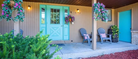 Une véranda en bois avec une porte turquoise, deux chaises, des jardinières suspendues et une petite table avec une chaise.