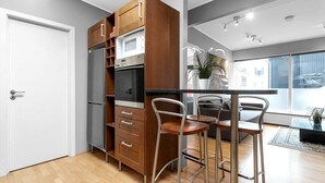 A modern kitchen with a built-in microwave, refrigerator, and wooden cabinets. There are two bar stools and a dining table.