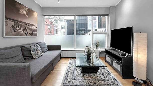 A modern living room with a grey sofa, a glass coffee table, a flat-screen TV, and a large window offering a view of the city.