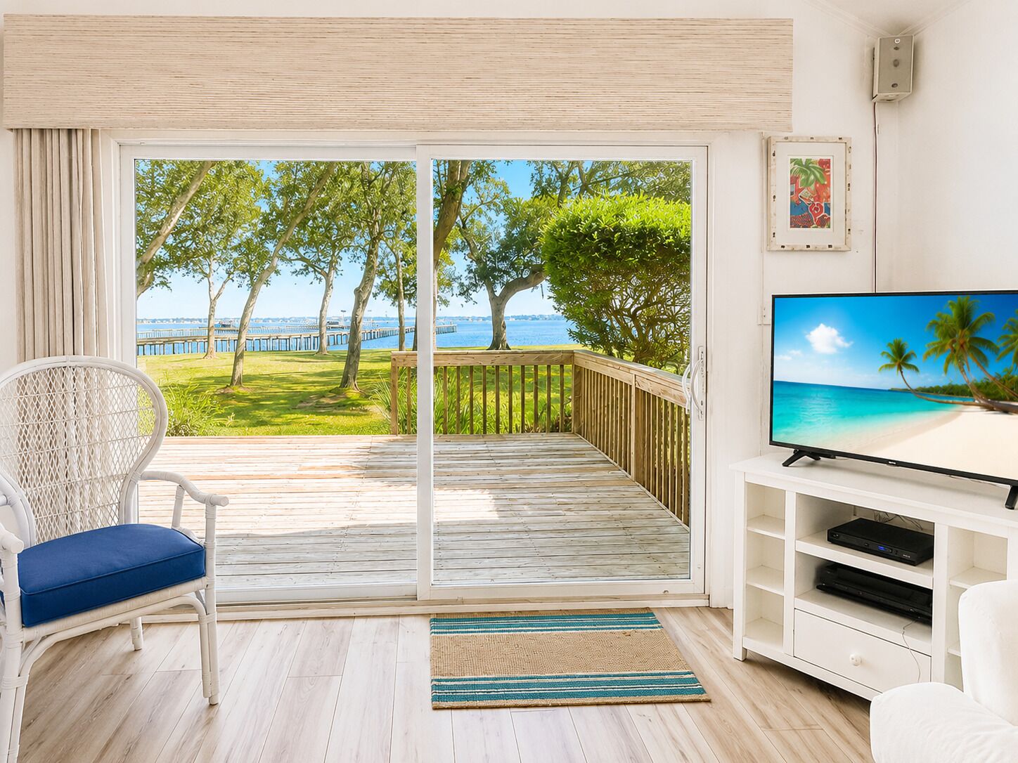 Ein Zimmer mit Fernseher auf einem weißen Schrank, ein Korbstuhl mit blauem Kissen, ein Holzterrasse mit Blick auf das Meer und Bäume sowie eine Glastür.