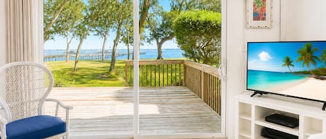 A room with a TV on a white stand, a wicker chair with a blue cushion, a wooden deck with a view of the sea and trees, and a sliding glass door.