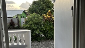 Blick durch eine Tür in einen Raum mit Fliesenboden, einer weißen Bank und einer Fußmatte. Davor befindet sich ein grünes Haus, ein Busch mit gelben Blüten und ein Kiesweg.