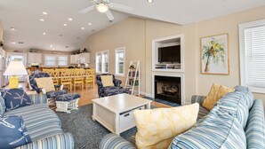 A spacious living room with a fireplace, a ceiling fan, and a dining area in the background. - Pirate's Booty: Private Pool | Hot Tub | Elevator (Nags Head)