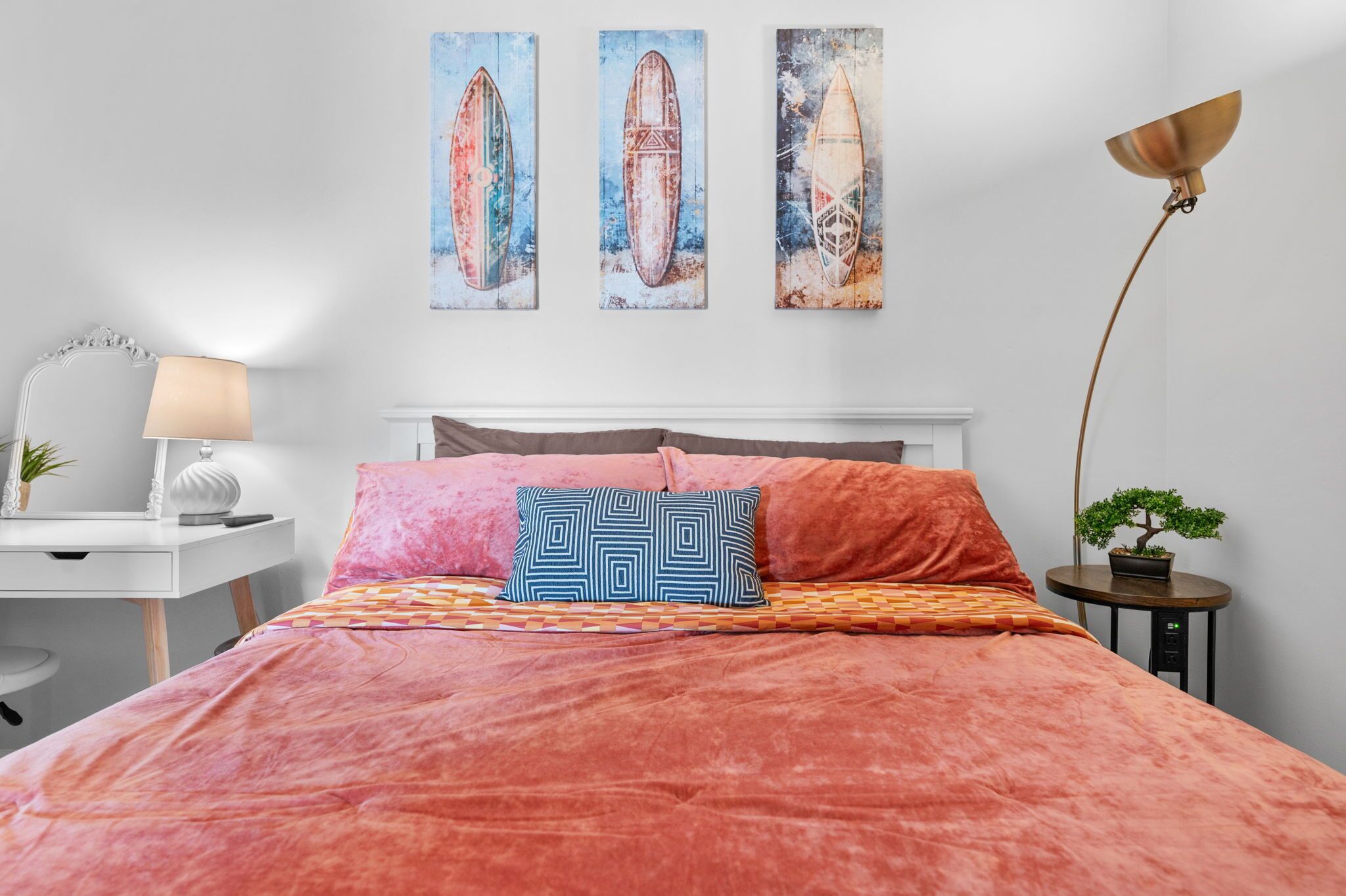 A bedroom with a large bed, a desk, a lamp, and three surfboard paintings on the wall.