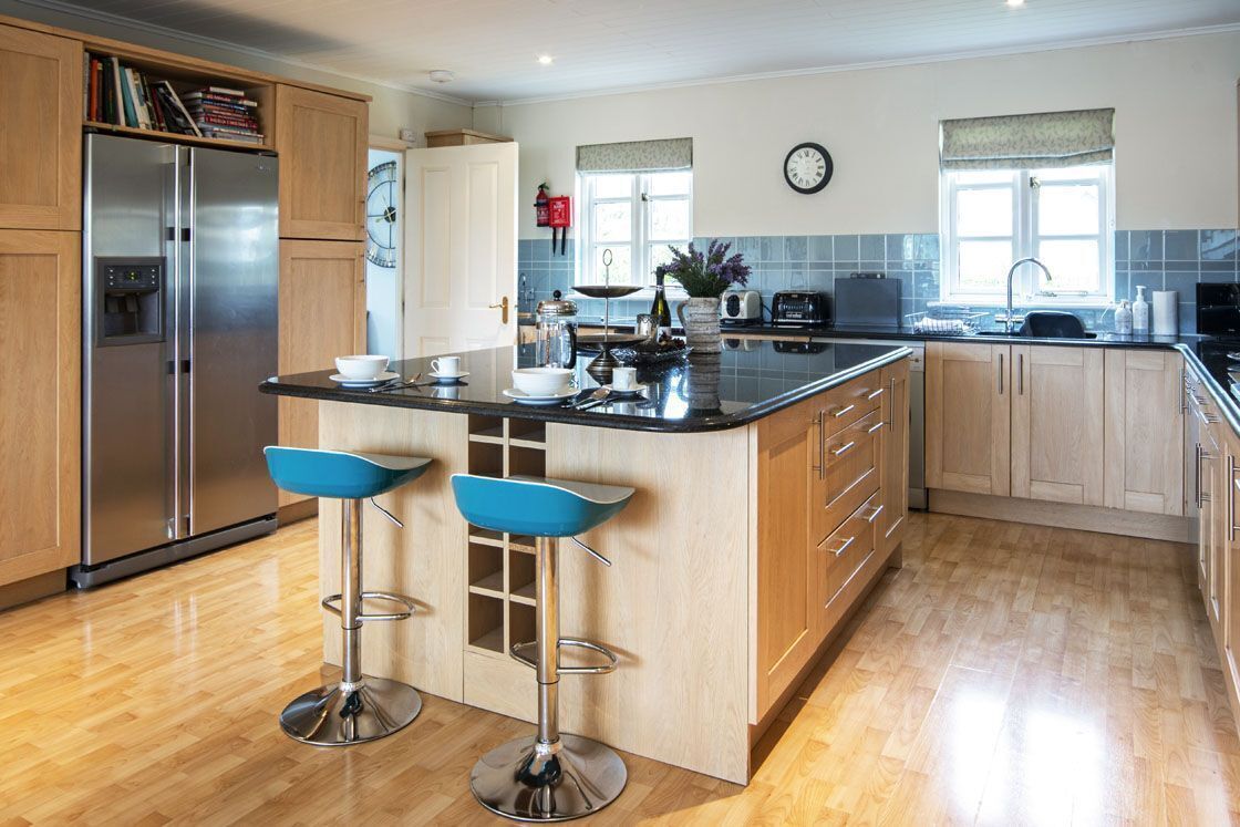A modern kitchen with a central island, two bar stools, wooden cabinets, a stainless steel refrigerator, and a clock on the wall.