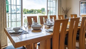 A dining table set for six with wooden chairs, large windows with curtains, and a view of greenery outside. - Windrush Lake 80/81, Waterlow Lodge (P) - Lakeside Lodge, Hot Tub, Sleeps 7 (South Cerney)