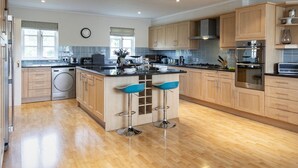 A modern kitchen with wooden cabinets, a central island with bar stools, a washing machine, and a clock on the wall. - Windrush Lake 80/81, Waterlow Lodge (P) - Lakeside Lodge, Hot Tub, Sleeps 7 (South Cerney)