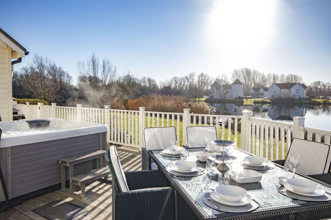 Un área de comedor al aire libre con jacuzzi, cerca blanca y vista a un lago y casas al fondo.