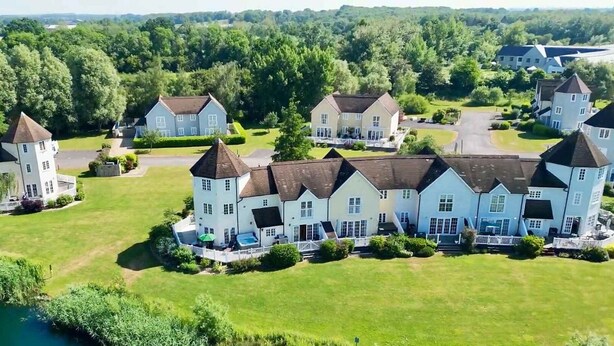 A large, multi-story building with a symmetrical facade, multiple chimneys, and a central courtyard with a swimming pool. - Windrush Lake 80/81, Waterlow Lodge (P) - Lakeside Lodge, Hot Tub, Sleeps 7 (South Cerney)