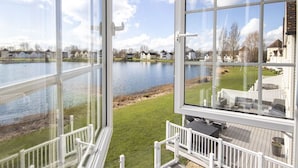 Une vue depuis une grande fenêtre donnant sur un lac, avec une terrasse et des meubles d’extérieur visibles.