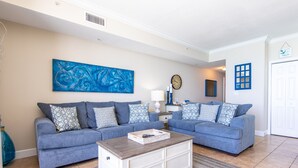 A living room with two blue sofas, a white coffee table, a blue abstract painting, a clock, and a blue-framed window.