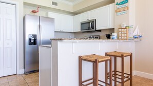 A kitchen with white cabinets, a stainless steel refrigerator, a microwave, and two wicker bar stools.