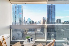 Un balcón con vista a la ciudad, una pequeña mesa con una planta y dos tazas, y una silla.