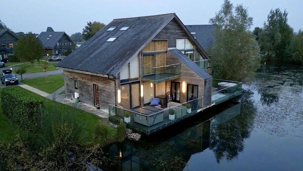 Et moderne hus med en glasbalcon der vender ud mod en flod, med en træfacade og et skråt tag.