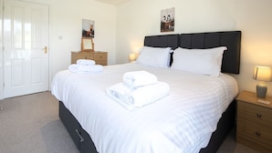 A neatly made bed with white linens and towels, a bedside table with a lamp, and a door in the background. - Windrush Lake 82, Puffin Lodge (P) - Great On-site Activities, Hot Tub, Sleeps 8 (SOUTH CERNEY)