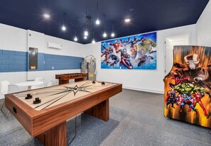 Een kamer met een tafelvoetbaltafel, een groot schilderij met superhelden en een deur met een gordijn in superhelden stijl.