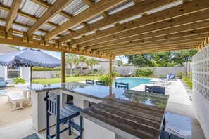 Ein Outdoor-Kochbereich mit Barbereich, Sitzgelegenheiten und Blick auf einen Hinterhof mit Schwimmbad.
