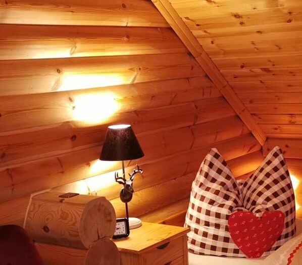 A cozy cabin room with wooden walls, a wooden nightstand with a lamp, a clock, and a checkered pillow with a heart-shaped pillow. - Chalet with a view of the castle - Vacation in the Harz Mountains, in Wernigerode (Chalet 3/3) (Wernigerode)