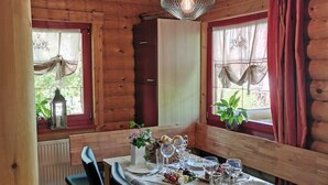 Interior de una cabaña de madera con una mesa de comedor preparada para cuatro personas, dos sillas, un armario y un colgador con luz.